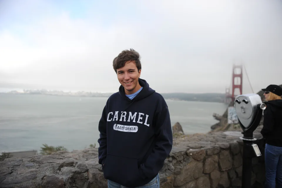 A young man at a scenic overlook with a red suspension bridge in the fog behind him