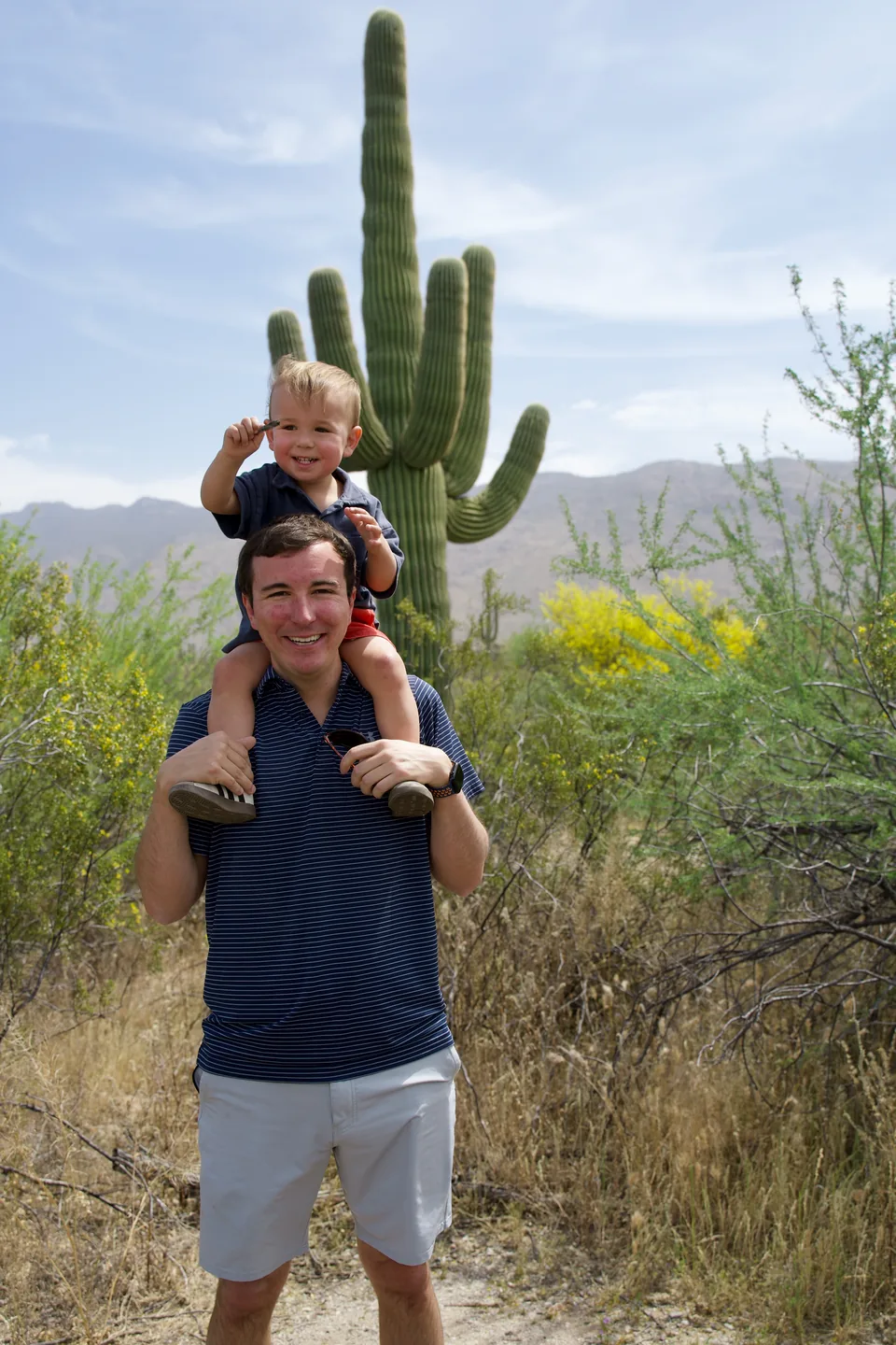 A father carrying a child on his shoulders on a desert trail beside a saguaro